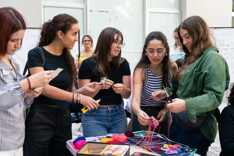 Grupo de mujeres jóvenes participa en una actividad colaborativa, manipulando hilos de colores alrededor de una mesa.