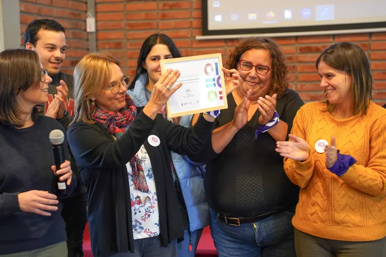 Grupo de personas aplaude mientras una integrante exhibe un reconocimiento en una actividad vinculada al Modelo de Calidad con Equidad de Género.