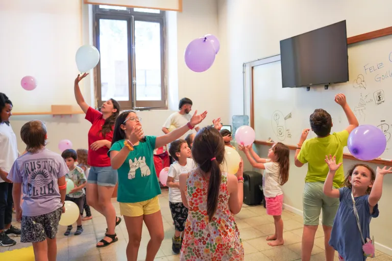 Grupo de niñas, niños y personas adultas participa en una actividad recreativa en un salón, jugando con globos de colores que lanzan al aire.
